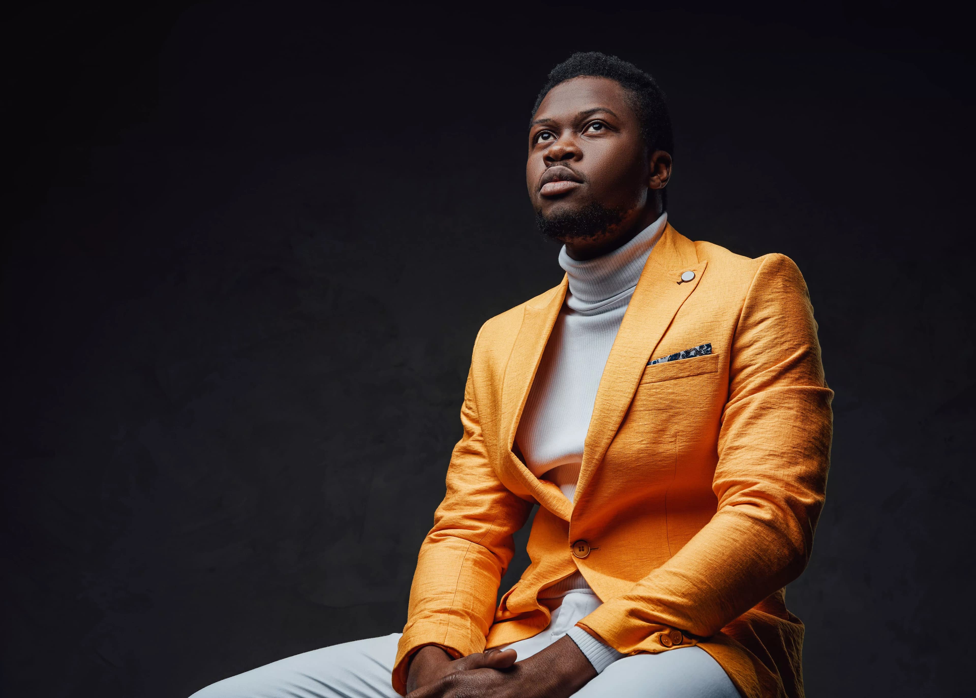 Un homme assis, veste ocre et col roulé blanc, le regard porté hors du cadre, sur fond sombre.